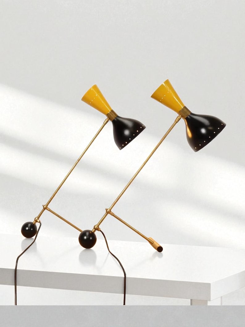 May include: Two mid-century modern desk lamps. Each lamp has a yellow conical shade, a black bell-shaped shade, and a brass-colored arm. The lamps are connected to a black base with a black sphere and a black cord.