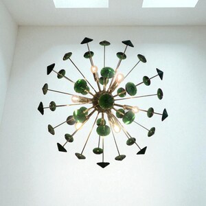May include: A modern chandelier with a central brass sphere, radiating outward with thin rods. Each rod features a green glass disc and a black triangle. The chandelier has several small lights and is mounted on a white ceiling.