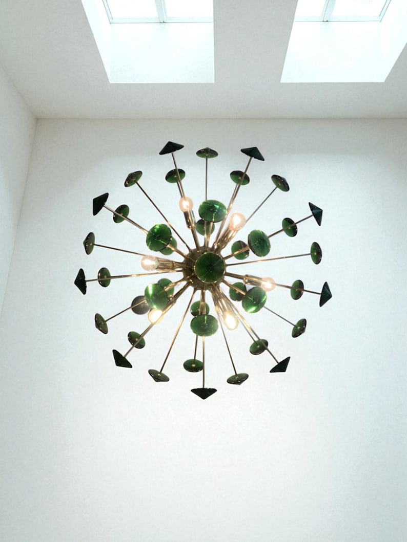 May include: A modern chandelier with a central brass sphere, radiating outward with thin rods. Each rod features a green glass disc and a black triangle. The chandelier has several small lights and is mounted on a white ceiling.