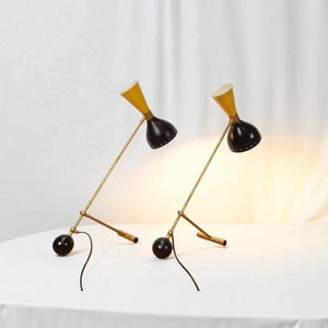 May include: Two mid-century modern desk lamps. Each lamp has a yellow conical shade, a black, bell-shaped head, and a brass-colored arm. The base is a black sphere. The lamps are on a white surface.