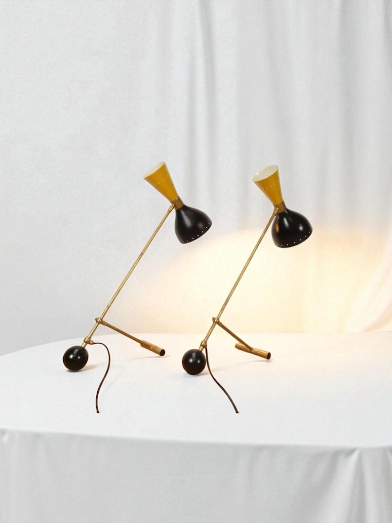 May include: Two mid-century modern desk lamps. Each lamp has a yellow conical shade, a black, bell-shaped head, and a brass-colored arm. The base is a black sphere. The lamps are on a white surface.