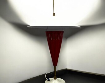 May include: A modern table lamp with a white lampshade and a red, cone-shaped base. The lamp has a brass stem and a white circular base. The design is minimalist and the lamp is set against a white wall.