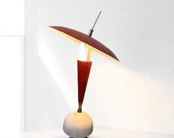 May include: A modern table lamp with a red shade and a white and gray base. The lamp has a conical red stem and a brass accent. The shade is a dark red color and is angled upwards. The lamp is on a white surface.