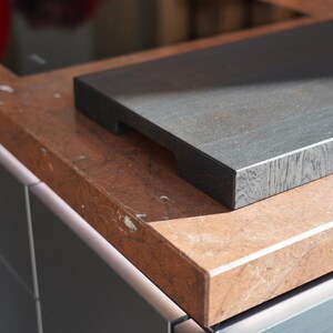 May include: A dark gray wooden cutting board with a smooth surface sits on a light brown countertop with a marbled pattern.