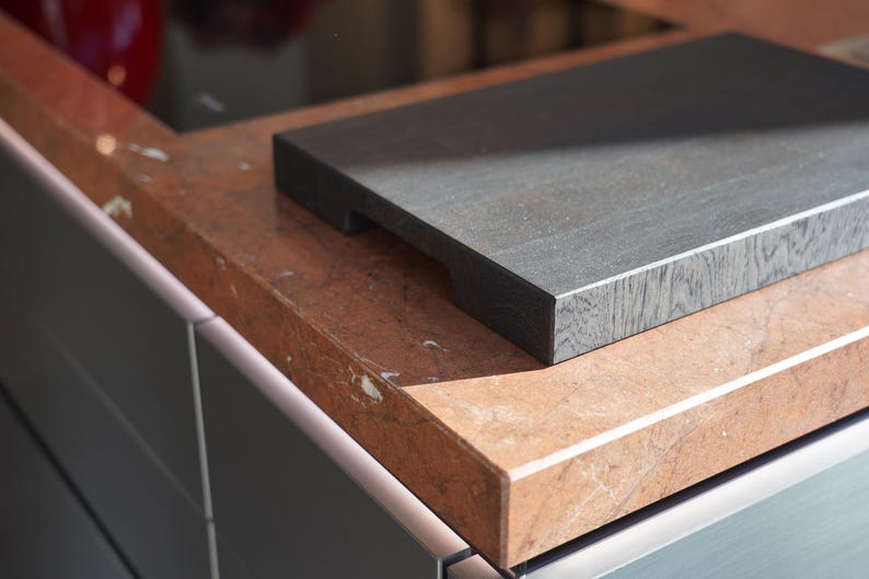 May include: A dark gray wooden cutting board with a smooth surface sits on a light brown countertop with a marbled pattern.