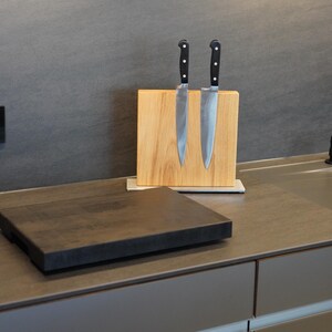 May include: Two silver kitchen knives with black handles are mounted on a wooden magnetic knife holder. The holder is on a gray countertop with a black cutting board in front of it.