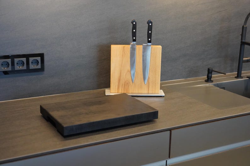 May include: Two silver kitchen knives with black handles are mounted on a wooden magnetic knife holder. The holder is on a gray countertop with a black cutting board in front of it.