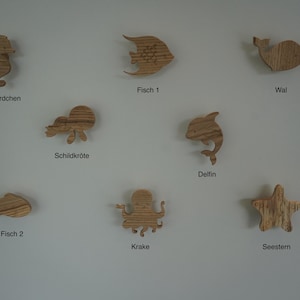 Könnte beinhalten: Eine Sammlung von Holzausschnitten von Meerestieren, darunter ein Seepferdchen, Fisch, Wal, Schildkröte, Delfin, Krake und Seestern. Jeder Ausschnitt besteht aus hellem Holz mit einem natürlichen Maserungsmuster. Deutsche Wörter darunter.
