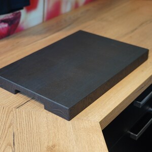 May include: A dark brown wooden cutting board with a smooth surface and rounded edges. The board is sitting on a light wood countertop.