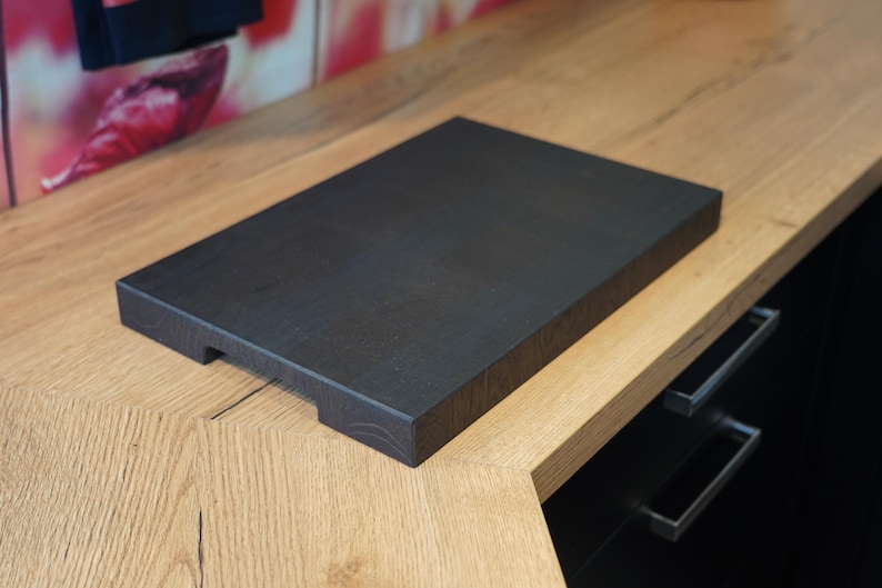 May include: A dark brown wooden cutting board with a smooth surface and rounded edges. The board is sitting on a light wood countertop.