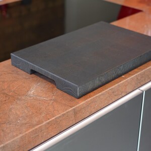 May include: A dark gray wooden cutting board with a handle on the side. The board is sitting on a brown countertop.