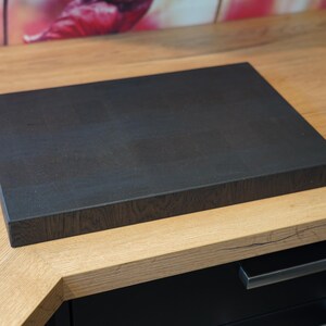 May include: A dark brown wooden cutting board with a textured surface. The board is sitting on a light brown wooden countertop.