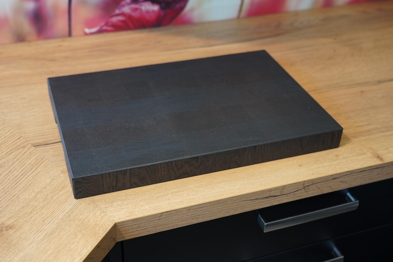 May include: A dark brown wooden cutting board with a textured surface. The board is sitting on a light brown wooden countertop.