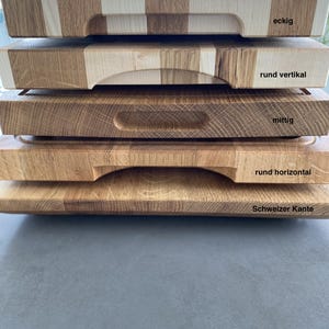 May include: Five wooden cutting boards stacked on top of each other. The top board has a square handle and the text "eckig". The second board has a rounded handle and the text "rund vertikal". The third board has a rectangular handle and the text "mittig". The fourth board has a rounded handle and the text "rund horizontal". The bottom board has a rounded edge and the text "Schweizer Kante".