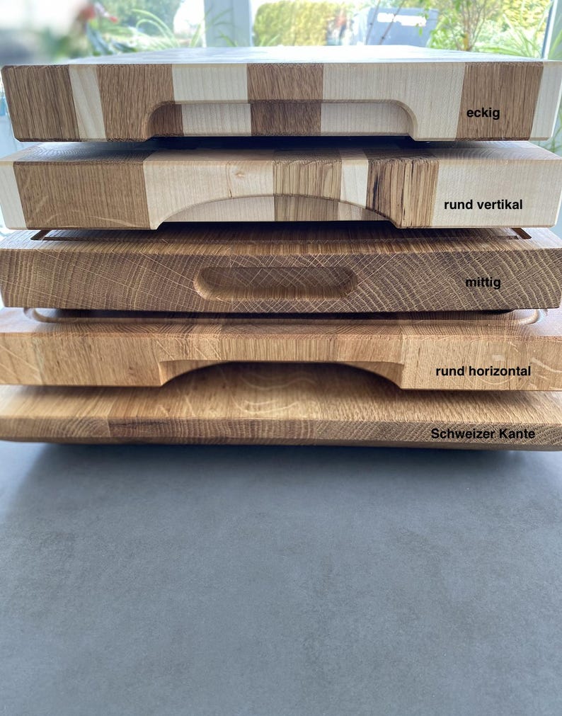 May include: Five wooden cutting boards stacked on top of each other. The top board has a square handle and the text "eckig". The second board has a rounded handle and the text "rund vertikal". The third board has a rectangular handle and the text "mittig". The fourth board has a rounded handle and the text "rund horizontal". The bottom board has a rounded edge and the text "Schweizer Kante".