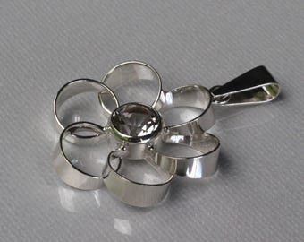 May include: A silver pendant shaped like a flower with six open, ribbon-like petals and a single, round, faceted clear gemstone set in the centre. The pendant includes a triangular bail at the top for attachment to a chain or necklace.