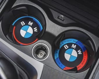 May include: Two black car coasters with the BMW logo in the center. The logo is a white, blue, and black design with the letters "B", "M", and "W" around the logo. The coasters are in a car's cup holders.
