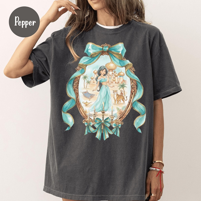 Puede incluir: Camiseta gris oscuro con una ilustraci&oacute;n enmarcada de una princesa con un vestido turquesa, con un tigre, un palacio y una alfombra m&aacute;gica. El marco est&aacute; adornado con lazos turquesas. La palabra "Pepper" est&aacute; en un c&iacute;rculo en la parte superior izquierda.