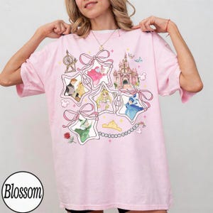 May include: Pink t-shirt featuring a star-shaped design with Disney princess illustrations, a castle, and bows. The shirt has short sleeves and a round neckline. The word "Blossom" is printed on the bottom left.