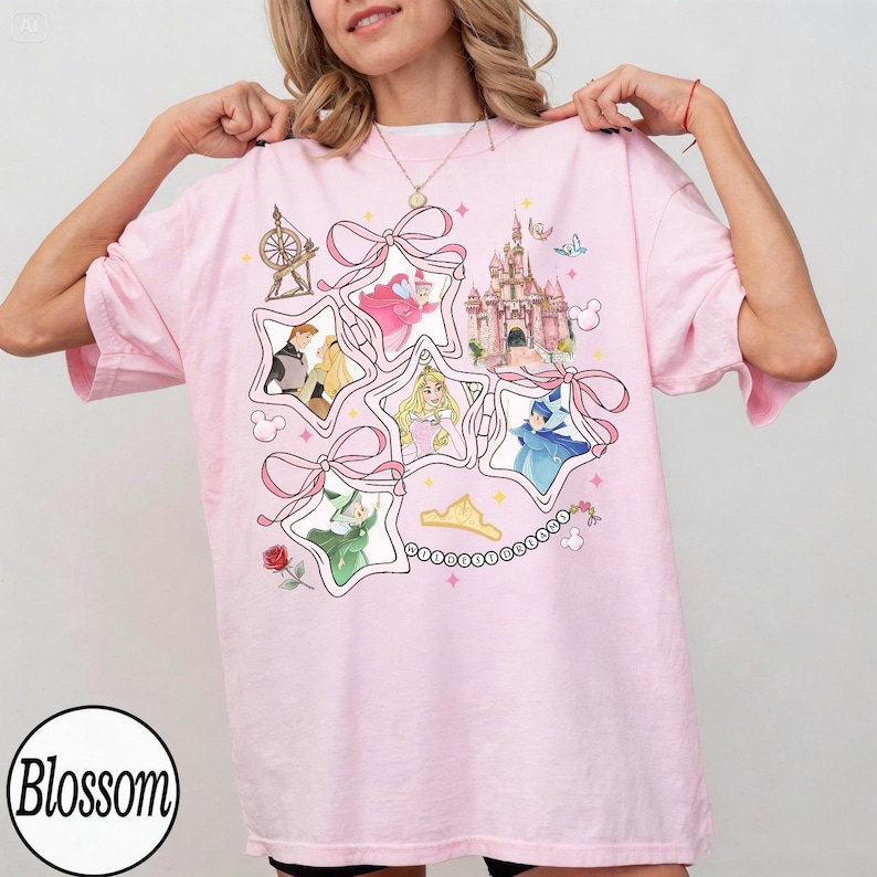 May include: Pink t-shirt featuring a star-shaped design with Disney princess illustrations, a castle, and bows. The shirt has short sleeves and a round neckline. The word "Blossom" is printed on the bottom left.