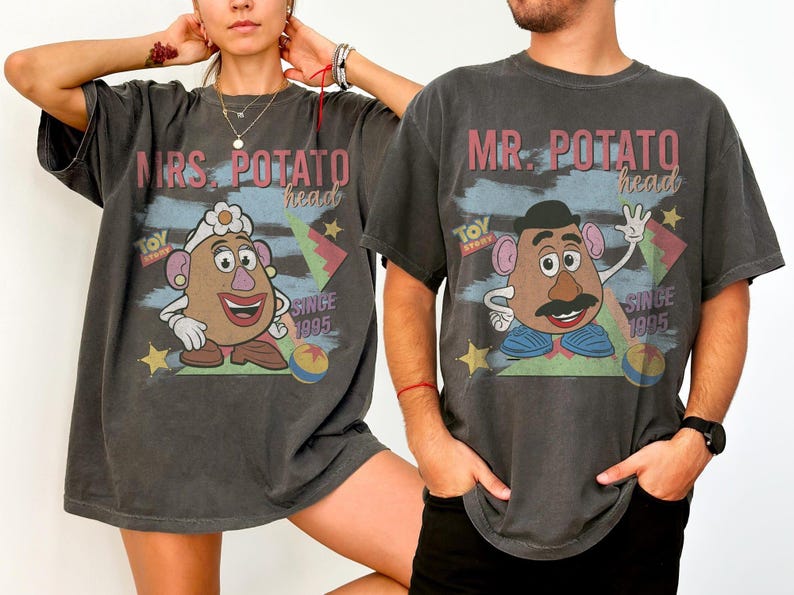 May include: Two charcoal gray t-shirts featuring Mr. and Mrs. Potato Head from Toy Story. The shirts have a distressed vintage look, with the characters' faces and the text "MRS. POTATO HEAD" and "MR. POTATO HEAD" printed on the front. The text "SINCE 1995" is also visible.
