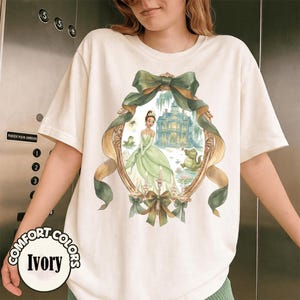 Puede incluir: Camiseta de color marfil con una ilustraci&oacute;n ovalada enmarcada en oro. La obra de arte representa a una princesa con un vestido verde, una mansi&oacute;n y una rana. El dise&ntilde;o est&aacute; acentuado con lazos y cintas verdes. Las palabras "COMFORT COLORS" e "Ivory" est&aacute;n impresas en la camiseta.