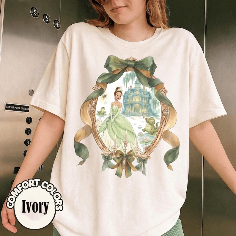 Puede incluir: Camiseta de color marfil con una ilustraci&oacute;n ovalada enmarcada en oro. La obra de arte representa a una princesa con un vestido verde, una mansi&oacute;n y una rana. El dise&ntilde;o est&aacute; acentuado con lazos y cintas verdes. Las palabras "COMFORT COLORS" e "Ivory" est&aacute;n impresas en la camiseta.