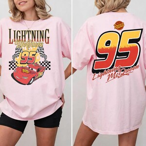 以下が含まれることがあります： ライトピンクのTシャツ。ライトニング・マックィーンと数字の95のグラフィックが特徴です。前面のデザインには「Lightning McQueen」の文字とチェッカーフラッグが含まれています。シャツの背面には大きな「95」と「Lightning McQueen」の文字が表示されています。