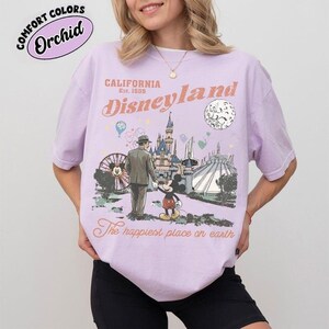 K&ouml;nnte beinhalten: Ein helllila T-Shirt mit einem Disneyland-Grafikdesign. Die Grafik zeigt eine Vintage-Illustration von Mickey Mouse, einem Mann und ikonischen Park-Wahrzeichen. Der Text "California Disneyland Est. 1955" und "The happiest place on earth" sind ebenfalls sichtbar.