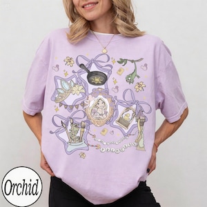 May include: A light purple t-shirt featuring a cartoon design with elements from a fairy tale. The design includes a crown, a frying pan, a portrait, and the text "The Lost Princess".