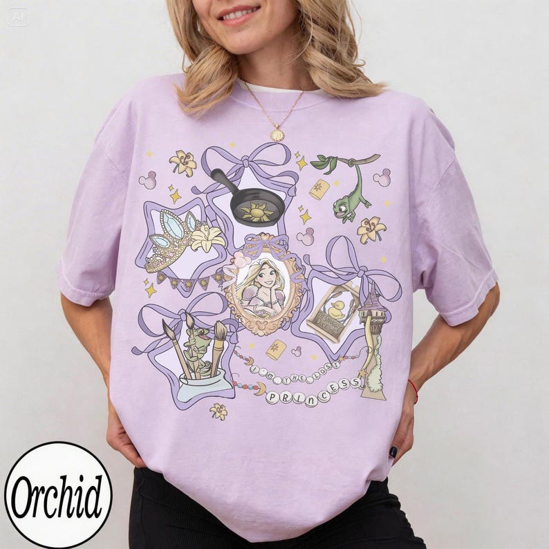 May include: A light purple t-shirt featuring a cartoon design with elements from a fairy tale. The design includes a crown, a frying pan, a portrait, and the text "The Lost Princess".