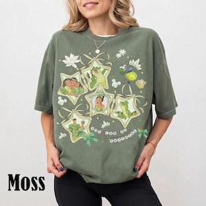 May include: Moss green t-shirt featuring a graphic design with cartoon characters in star-shaped frames. The design includes the text "You Can Do Anything". The shirt has short sleeves and a relaxed fit.