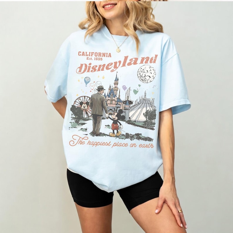 K&ouml;nnte beinhalten: Hellblaues T-Shirt mit Disneyland-Grafik. Die Grafik zeigt eine Illustration im Vintage-Stil von Mickey Mouse, einem Mann und Disneyland-Wahrzeichen. Der Text auf dem Shirt lautet "CALIFORNIA Est. 1955 Disneyland" und "The happiest place on earth."