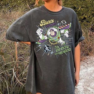 Pu&ograve; includere: Una t-shirt a maniche corte color grigio antracite con una grafica di Buzz Lightyear in volo. Il design include la scritta "Buzz Lightyear" e "To Infinity and Beyond" con accenti a forma di stella e un piccolo logo da ranger spaziale.