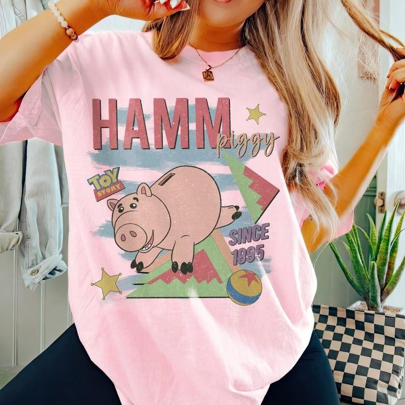 May include: Pink t-shirt featuring a graphic of Hamm from Toy Story. The design includes the text "HAMM piggy" and "SINCE 1995". The cartoon pig is depicted in a playful pose with a blue, green, and red background.