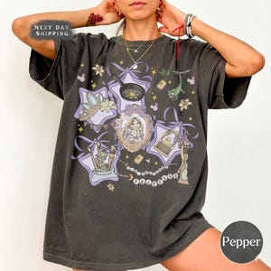May include: Dark gray t-shirt featuring a Rapunzel-themed design with purple accents. The graphic includes a portrait of Rapunzel, a crown, and other elements from the movie. The word "Princess" is spelled out in a beaded necklace design.