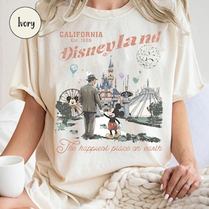 K&ouml;nnte beinhalten: Elfenbeinfarbenes T-Shirt mit Disneyland-Grafik. Das Design zeigt die Worte "California Disneyland Est. 1955" und eine Illustration von Mickey Mouse, einem Mann und Wahrzeichen des Parks. Der Text "The happiest place on earth" ist ebenfalls enthalten.