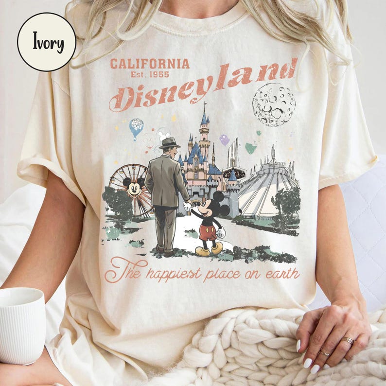 K&ouml;nnte beinhalten: Elfenbeinfarbenes T-Shirt mit Disneyland-Grafik. Das Design zeigt die Worte "California Disneyland Est. 1955" und eine Illustration von Mickey Mouse, einem Mann und Wahrzeichen des Parks. Der Text "The happiest place on earth" ist ebenfalls enthalten.
