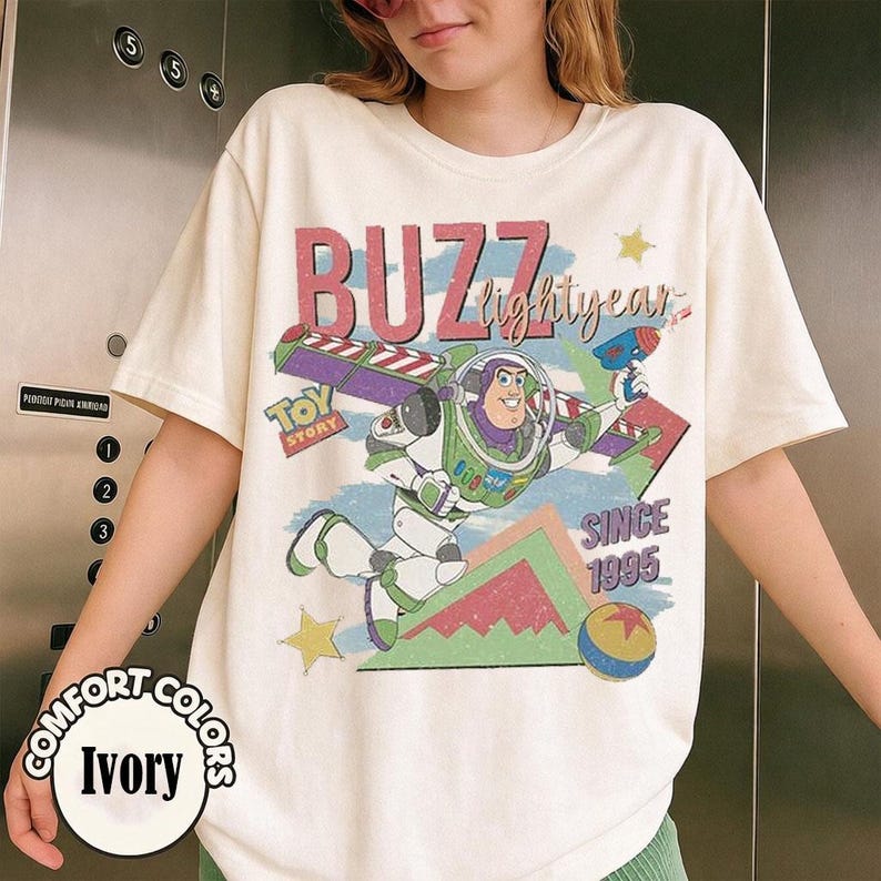 May include: Ivory-colored t-shirt featuring a graphic of Buzz Lightyear from Toy Story. The design includes the text "BUZZ lightyear" and "SINCE 1995". The shirt also has the words "COMFORT COLORS" and "Ivory" printed on it.