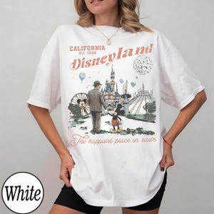 K&ouml;nnte beinhalten: Wei&szlig;es T-Shirt mit einem Vintage-Disneyland-Motiv. Das Design enth&auml;lt den Text "California Disneyland Est. 1955" und eine Illustration von Mickey Mouse, einem Mann und dem Schloss. Der Satz "The happiest place on earth" ist ebenfalls vorhanden.