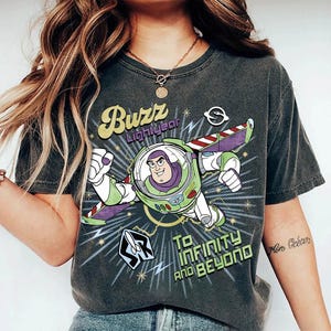 Pu&ograve; includere: Una t-shirt a maniche corte grigio antracite con una grafica di Buzz Lightyear in volo. Il design include il testo "Buzz Lightyear" e "To Infinity and Beyond" in un carattere retr&ograve;, circondato da stelle, fulmini e una piccola icona a forma di razzo.