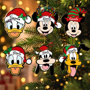 May include: Six flat acrylic Christmas ornaments featuring cartoon character faces including Daisy Duck, Mickey Mouse, Minnie Mouse, Donald Duck, Goofy, and Pluto. Each character wears a holiday hat with the names Sophia or David printed on them.