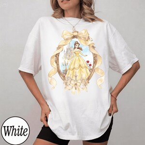 Puede incluir: Camiseta blanca con una ilustraci&oacute;n en acuarela de una mujer con un vestido amarillo dentro de un marco dorado con un lazo. El marco incluye un castillo y rosas rojas. La palabra "White" est&aacute; en un c&iacute;rculo blanco en la parte inferior.