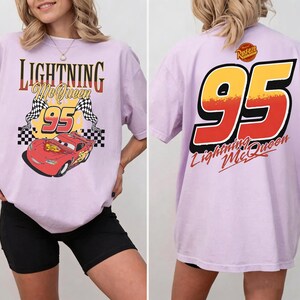 以下が含まれることがあります： ライトニング・マックィーンのグラフィックが特徴の薄紫色のTシャツ。前面には、赤いレーシングカーとチェッカーフラッグとともに「Lightning McQueen 95」の文字がプリントされています。背面には「95」の大きな数字と「Lightning McQueen」の文字がプリントされています。