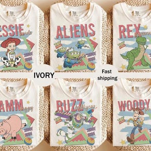 May include: Ivory-colored t-shirts featuring various Toy Story characters: Jessie, Aliens, Rex, Hamm, Buzz, and Woody. Each shirt has a character graphic with the text "Toy Story" and "Since 1995". The shirts are displayed on a woven surface.
