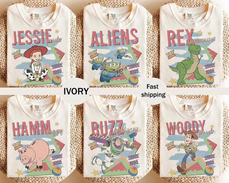 May include: Ivory-colored t-shirts featuring various Toy Story characters: Jessie, Aliens, Rex, Hamm, Buzz, and Woody. Each shirt has a character graphic with the text "Toy Story" and "Since 1995". The shirts are displayed on a woven surface.