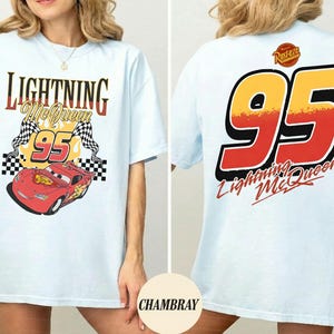 以下が含まれることがあります： ライトブルーのTシャツ。前面には「Lightning McQueen 95」の文字と赤いレーシングカー、チェッカーフラッグが描かれています。背面には、赤と黄色の大きな「95」と「Lightning McQueen」の文字がプリントされています。