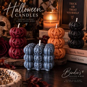 May include: A collection of wax candles shaped like stacked jack-o'-lanterns in deep red, orange, black, and a blue-grey cube formation. Each candle features carved pumpkin faces with a single white wick protruding from the top of the uppermost pumpkin.