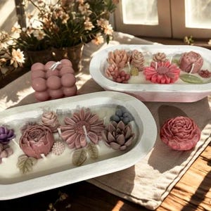 May include: A collection of decorative candles in various shapes and colors. The image features a bubble candle, floral candles, and a single rose-shaped candle. The candles are arranged on a neutral-colored surface, with a lit candle in a glass jar in the background.