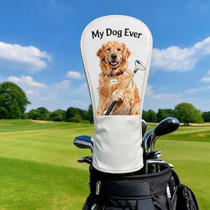 Könnte beinhalten: Eine weiße Golfschlägerhaube mit einer Illustration eines Golden Retrievers, einem Golfschläger und dem Text My Dog Ever. Die Haube steckt auf einem Schläger in einer schwarzen Golftasche vor einem grünen Golfplatz unter blauem Himmel.
